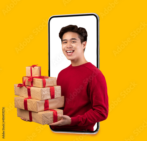 Smiling millennial asian man with many gift boxes on blank screen of big phone isolated on orange background, studio © Prostock-studio