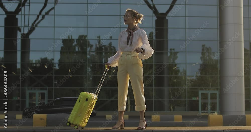 Slim young caucasian business woman looks at her wristwatch, looking around as she walks near airport. Blonde is carrying yellow suitcase in strange city. Travel and lifestyle concept. Slim young caucasian business woman looks at her wristwatch, looking around as she walks near airport. Blonde is carrying yellow suitcase in strange city. Travel and lifestyle concept.