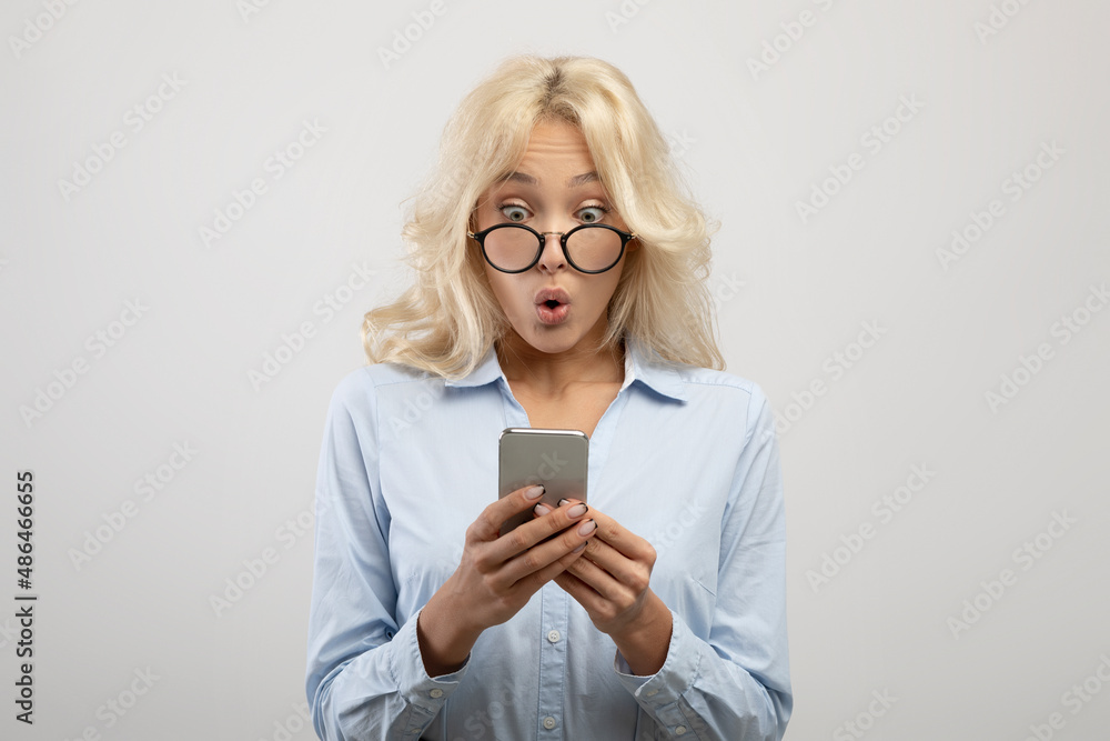 Young shocked businesswoman in glasses holding and looking at smartphone, shouting OMG, standing over light background