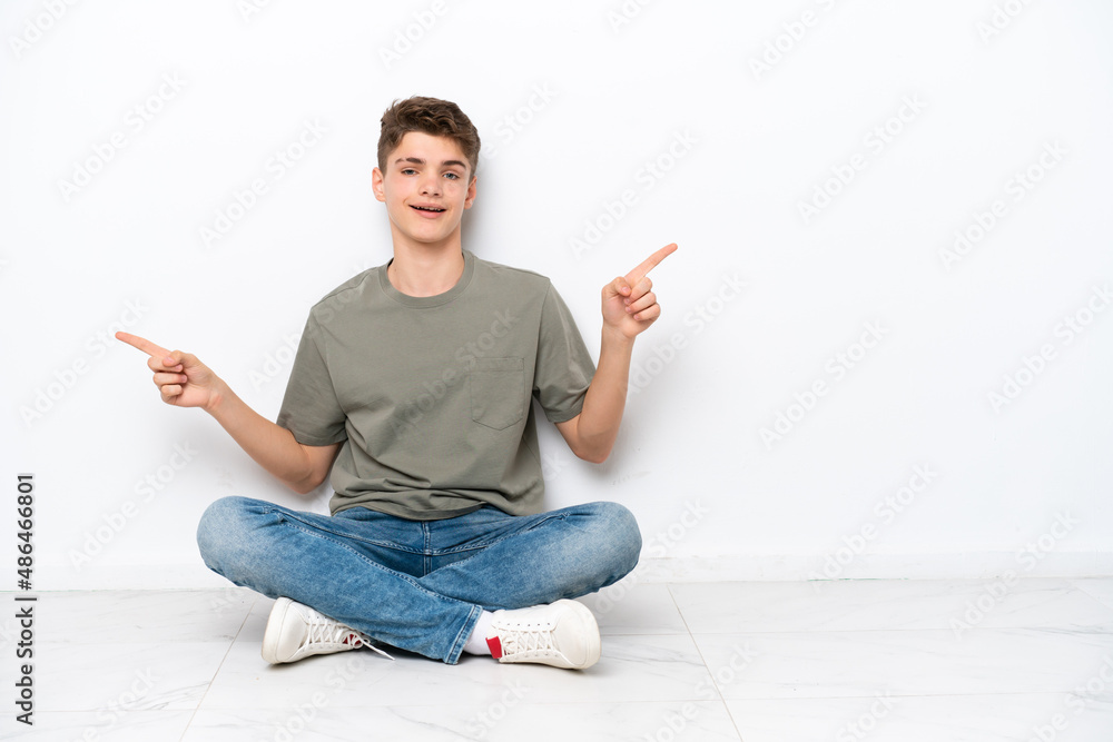 Teenager Russian man sitting on the floor isolated on white background pointing finger to the laterals and happy Teenager Russian man sitting on the floor isolated on white background pointing finger to the laterals and happy