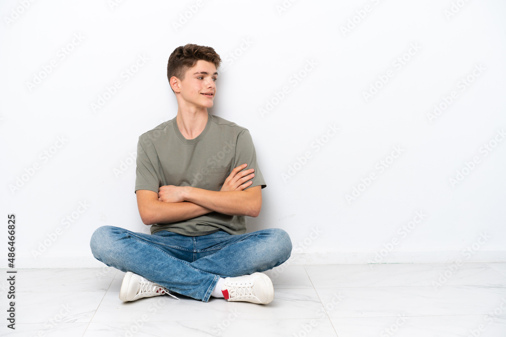 Teenager Russian man sitting on the floor isolated on white background with arms crossed and happy Teenager Russian man sitting on the floor isolated on white background with arms crossed and happy