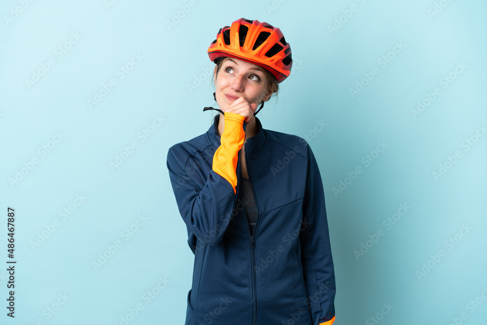 Young cyclist woman isolated on blue background and looking up - obrazy, fototapety, plakaty 