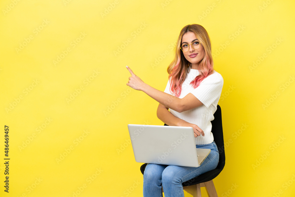 Young woman sitting on a chair with laptop over isolated yellow background pointing back - obrazy, fototapety, plakaty 