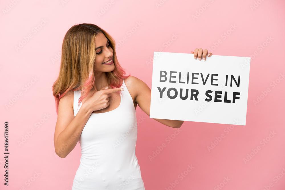Young woman over isolated pink background holding a placard with text Believe In Your Self and pointing it Young woman over isolated pink background holding a placard with text Believe In Your Self and pointing it