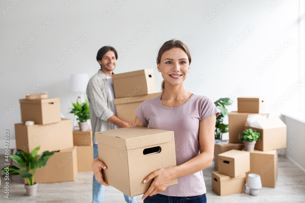 Smiling millennial caucasian lady and guy in casual clothes carry cardboard boxes with belongings in room interior Smiling millennial caucasian lady and guy in casual clothes carry cardboard boxes with belongings in room interior