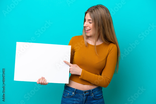 Young caucasian woman isolated on blue background holding an empty placard with happy expression and pointing it