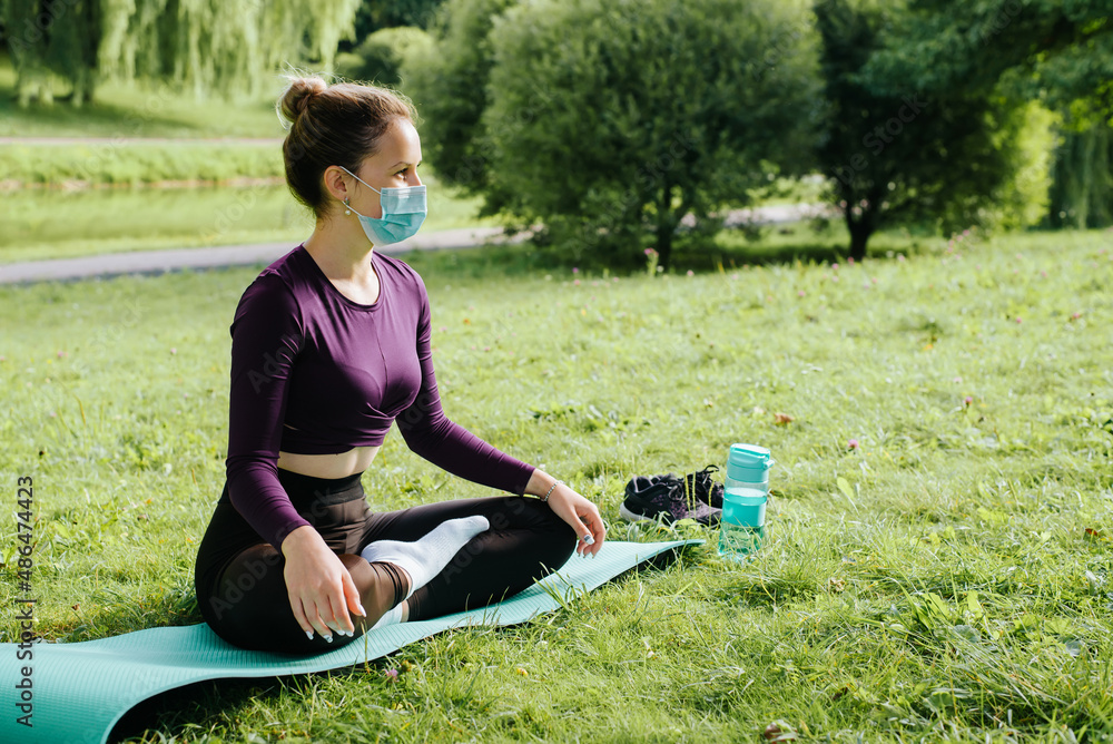 Side view of young woman in medical protective face mask practicing yoga and meditation sitting in lotus position on mat on green grass in park. Sportswoman exercising outdoors. New normal Side view of young woman in medical protective face mask practicing yoga and meditation sitting in lotus position on mat on green grass in park. Sportswoman exercising outdoors. New normal