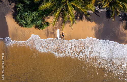 Canvastavla Aerial photo of surfer lying next to surfboard on the sandy ocean shore in STAR pose and sun tanning under tall palm trees