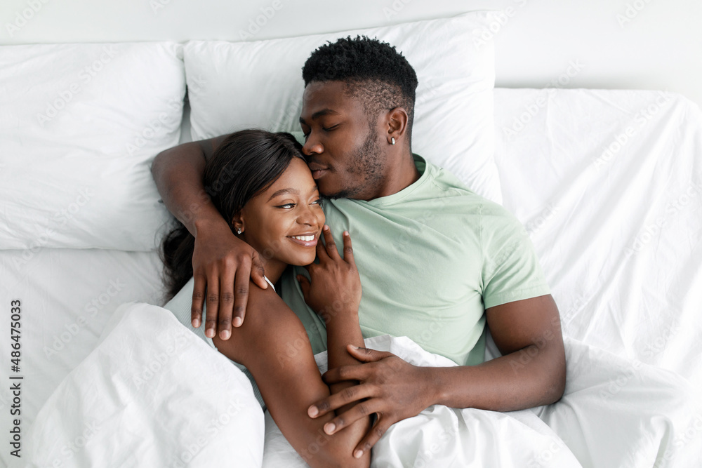 Cheerful young african american husband hugging wife and kissing on comfortable bed, resting, relax in bedroom