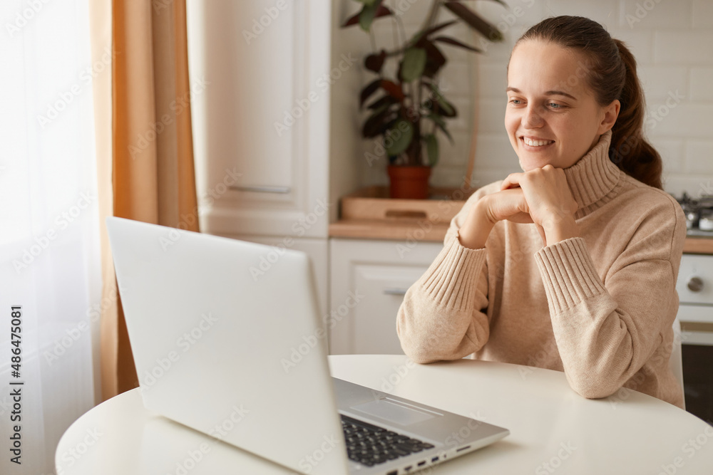 Indoor shot of smiling woman wearing beige sweater sitting in kitchen and having online lesson on laptop, looking at notebook screen with pleasant smile, having video call. Indoor shot of smiling woman wearing beige sweater sitting in kitchen and having online lesson on laptop, looking at notebook screen with pleasant smile, having video call.