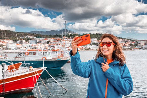 a woman in a jacket takes a selfie photo on a smartphone against the background of a pier with fishing boats photo