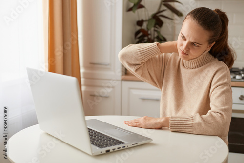 Portrait of tired woman wearing beige jumper sitting in kitchen and working online on laptop, being exhausted, feeling pain in neck, touching and massaging shoulders, looking at computer display.