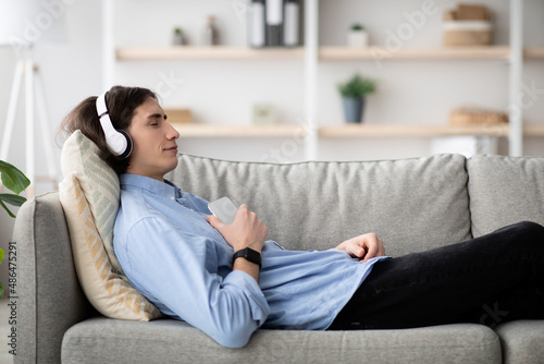 Rest and relax concept. Calm guy lying on sofa, listening to music, enjoying meditation for sleep in wireless headphones