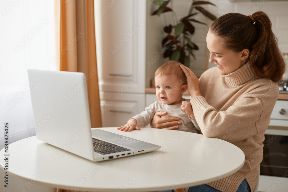 Indoor shot of mother wearing beige sweater sitting in kitchen, working online on laptop and holding her infant daughter, having freelance work during maternity leave. Indoor shot of mother wearing beige sweater sitting in kitchen, working online on laptop and holding her infant daughter, having freelance work during maternity leave.