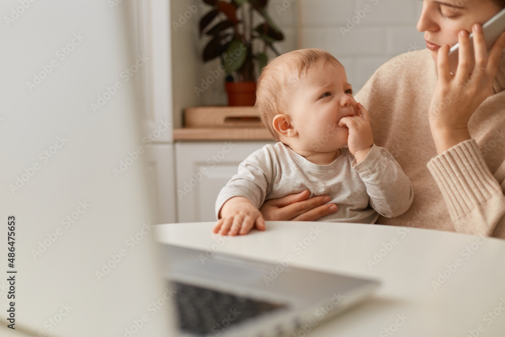 Cropped image woman wearing beige sweater sitting in kitchen and working online on laptop and talking via smartphone, looking after baby girl, calming down crying daughter. Cropped image woman wearing beige sweater sitting in kitchen and working online on laptop and talking via smartphone, looking after baby girl, calming down crying daughter.