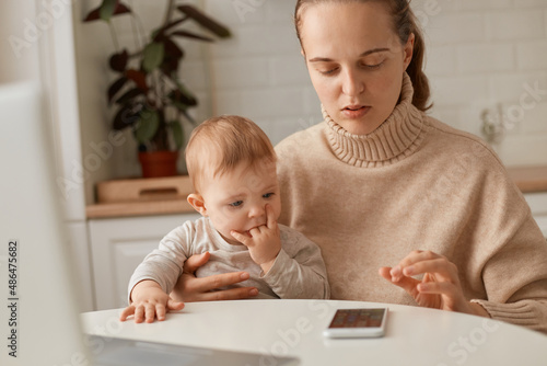 Indoor shot of concentrated serious woman wearing beige sweater sitting in kitchen and working online on her mobile phone, combines her work and maternity leave.