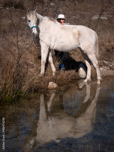 Female horse owner with white hat and her horse looking curiously to one side  reflection in water.