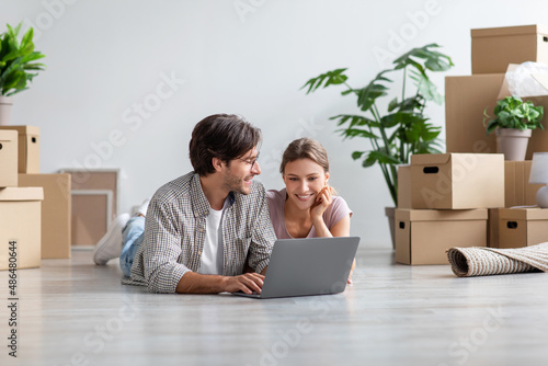 Pleased european millennial couple lie on floor look at computer among cardboard boxes with stuff in room