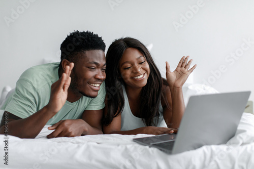 Cheerful young african american wife and husband lie on bed waving hand at computer webcam in bedroom