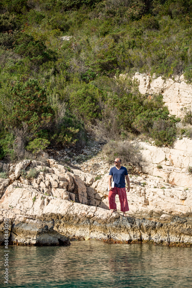 Digitaler Nomade mit Sonnenbrille, roter Hose und Laptop unterm Arm steht auf Felsen am Meer und blickt in die Ferne, Hochformat