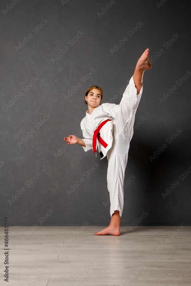 young girl karateka in a white kimono and a red belt trains and performs a set of exercises against a gray wall