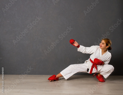young karateka girl in a white kimono and red competition outfit trains and performs a set of exercises against a gray wall