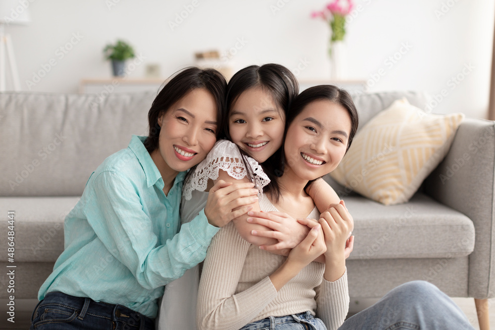 Young Asian woman with her cute daughter and mature mother hugging and smiling at camera indoors Young Asian woman with her cute daughter and mature mother hugging and smiling at camera indoors