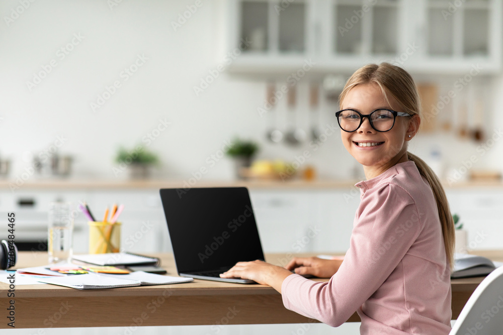 Cheerful caucasian teen schoolgirl in glasses look at camera at table with computer with blank screen Cheerful caucasian teen schoolgirl in glasses look at camera at table with computer with blank screen