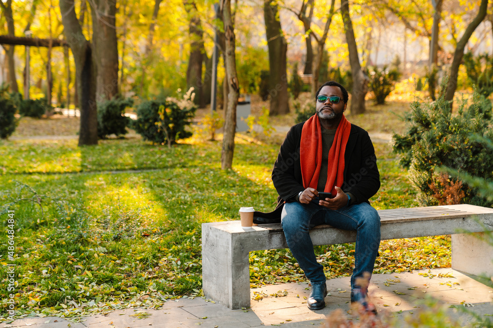 Fototapeta premium Black bearded man in sunglasses using cellphone while sitting on bench