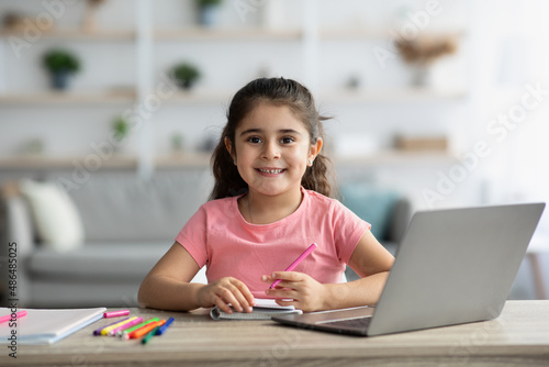 Cute Little Girl Sitting At Table With Laptop And Drawing In Notepad