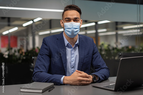 Arab manager in face mask sitting at worktable with computer