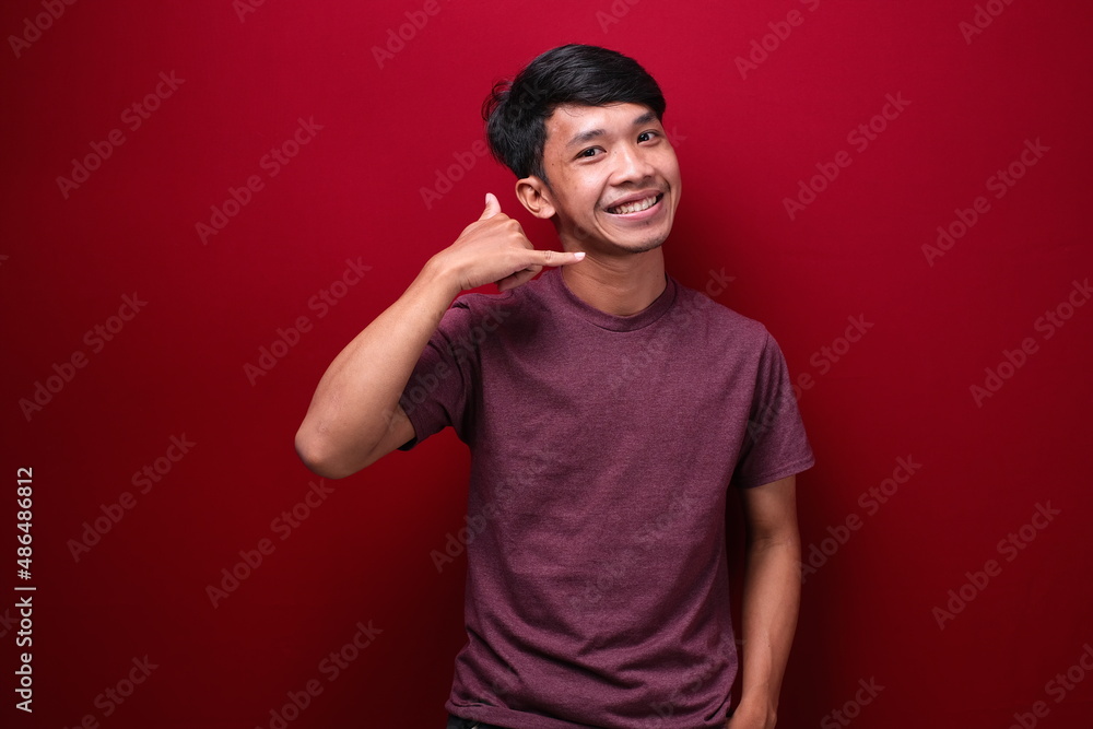 Young asian man red background smiling doing phone gesture with hand and fingers like talking on the telephone. Communication concepts. Young asian man red background smiling doing phone gesture with hand and fingers like talking on the telephone. Communication concepts.