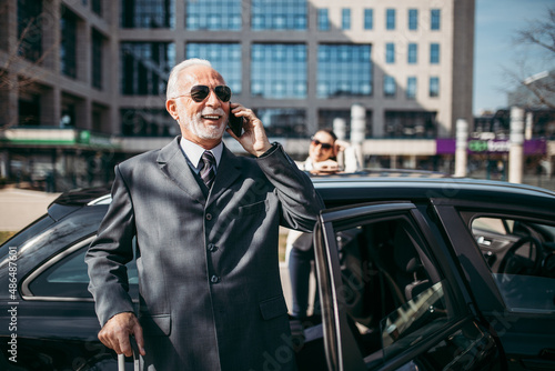Good looking senior business man and his young woman colleague or coworker standing by the car. He talking on smartphone, holds a suitcase and prepares to go on a business trip.  © Dusko