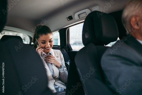 Good looking young business woman sitting on backseat in luxury car. She using her smart phone and smiling. Transportation in corporate business concept. © Dusko