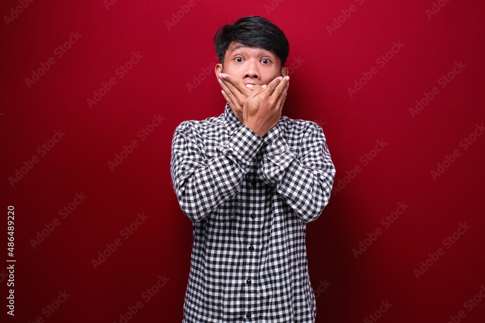 asian man wearing plaid shirt with shocked expression, on red background asian man wearing plaid shirt with shocked expression, on red background
