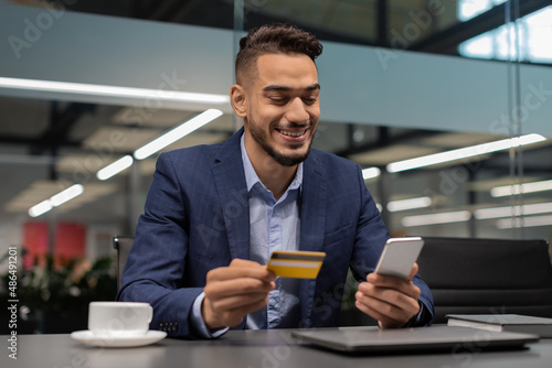 Cheerful middle eastern entrepreneur using smartphone, holding credit card