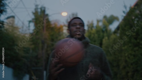 Portrait of real authentic millennial Black man walk on street towards camera at dusk, toss basketball from hand to hand. Cinematic shot of athletic youth on early morning training