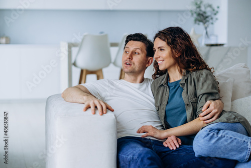 Happy newlyweds with dark hair sit on comfortable sofa in living room and discuss plans for future smiling broadly closeup