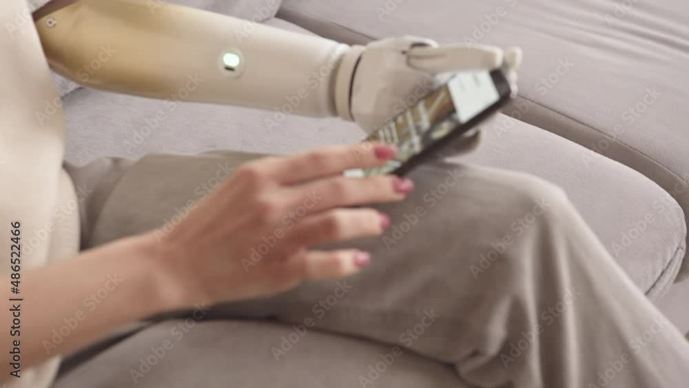 Slowmo shot of young Caucasian woman with bionic arm using food delivery app on her smartphone sitting on sofa at home Slowmo shot of young Caucasian woman with bionic arm using food delivery app on her smartphone sitting on sofa at home