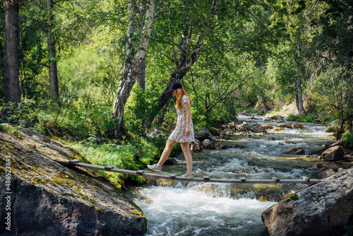 Young woman in a summer dress walking on a wooden bridge over mountain stream. Excursion in nature reserve. Picturesque forest landscape.
