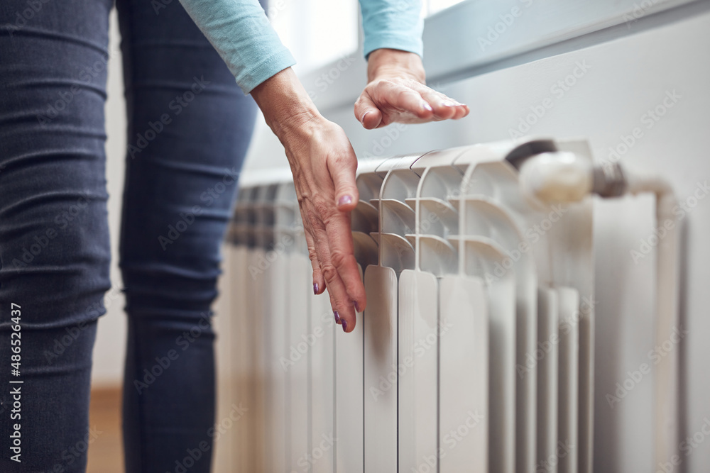 Woman heating her hands on the radiator during cold winter days. - obrazy, fototapety, plakaty 