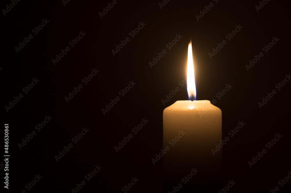 A single burning candle flame or light glowing on a white candle on black or dark background on table in church for Christmas, funeral or memorial service A single burning candle flame or light glowing on a white candle on black or dark background on table in church for Christmas, funeral or memorial service