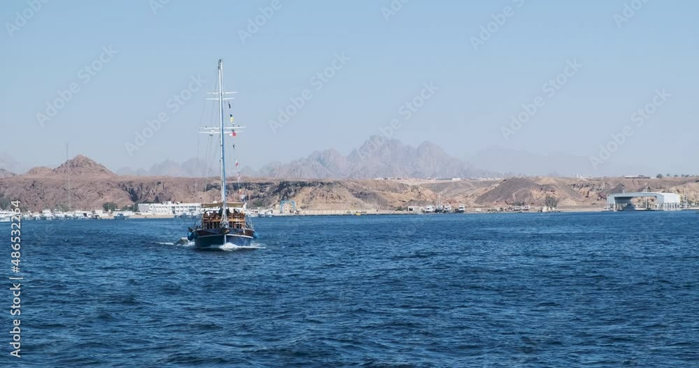 An old wooden ship, bow view, with lowered sails, emerges from the bay. View of the harbor in Sharm El Sheikh, desert, sand, pier. An old wooden ship, bow view, with lowered sails, emerges from the bay. View of the harbor in Sharm El Sheikh, desert, sand, pier.
