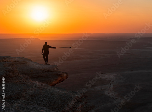 Man with a camera on a selfie stick walking along a cliff at sunset, The Edge of the World (Jebel Fihrayn), Saudi Arabia photo