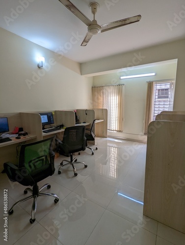 A big office interior with a few wooden desks with computer and black chairs in front of each placed in a row, with a ceiling fan, lights, big window with curtain and a shiny floor on the surrounding. photo