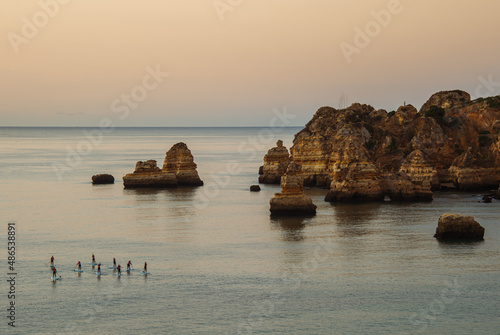SUP touring. Group of tourists on sap boards slowly swim on smooth water to explore local unusual rocks from water. Lagos, Portugal