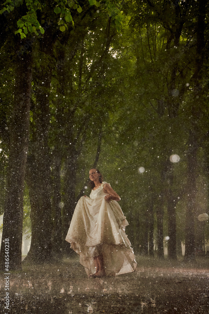 happy bride in a white wedding dress in the rain on an alley under big green trees walking peaying sitting and laughing