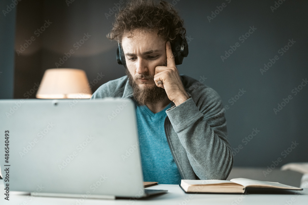 Serious young freelancer man working at laptop sitting at the desk at home office and taking notes. Remote working pensive young man working at home. Work from home concept Serious young freelancer man working at laptop sitting at the desk at home office and taking notes. Remote working pensive young man working at home. Work from home concept