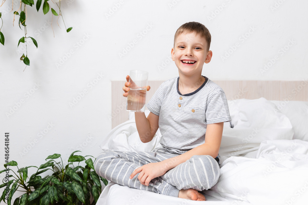 Health concept. Happy active Child smiling, when hold fresh pure water in a glass siting on bed in morning. Taking care of your body. Awakening Ritual Health concept. Happy active Child smiling, when hold fresh pure water in a glass siting on bed in morning. Taking care of your body. Awakening Ritual
