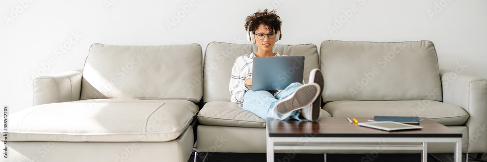 Young curly woman in headphones using laptop while sitting on couch Young curly woman in headphones using laptop while sitting on couch